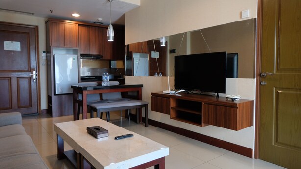 A modern living room with a TV, a dining table, and a kitchen area with wooden cabinets and a refrigerator. - Homey 2Br At Galeri Ciumbuleuit 1 Apartment (Bandung)