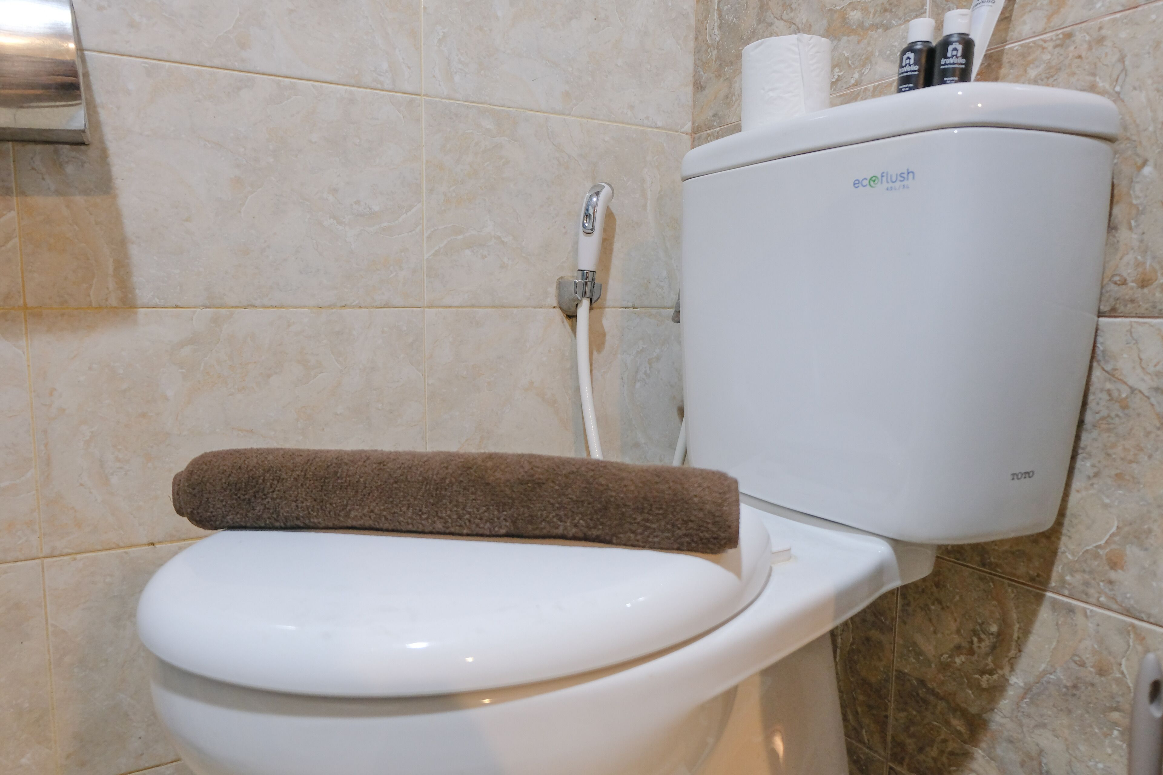 Een toilet met een bruine handdoek op de zitting, een toiletrolhouder en een waterfles op de tank.