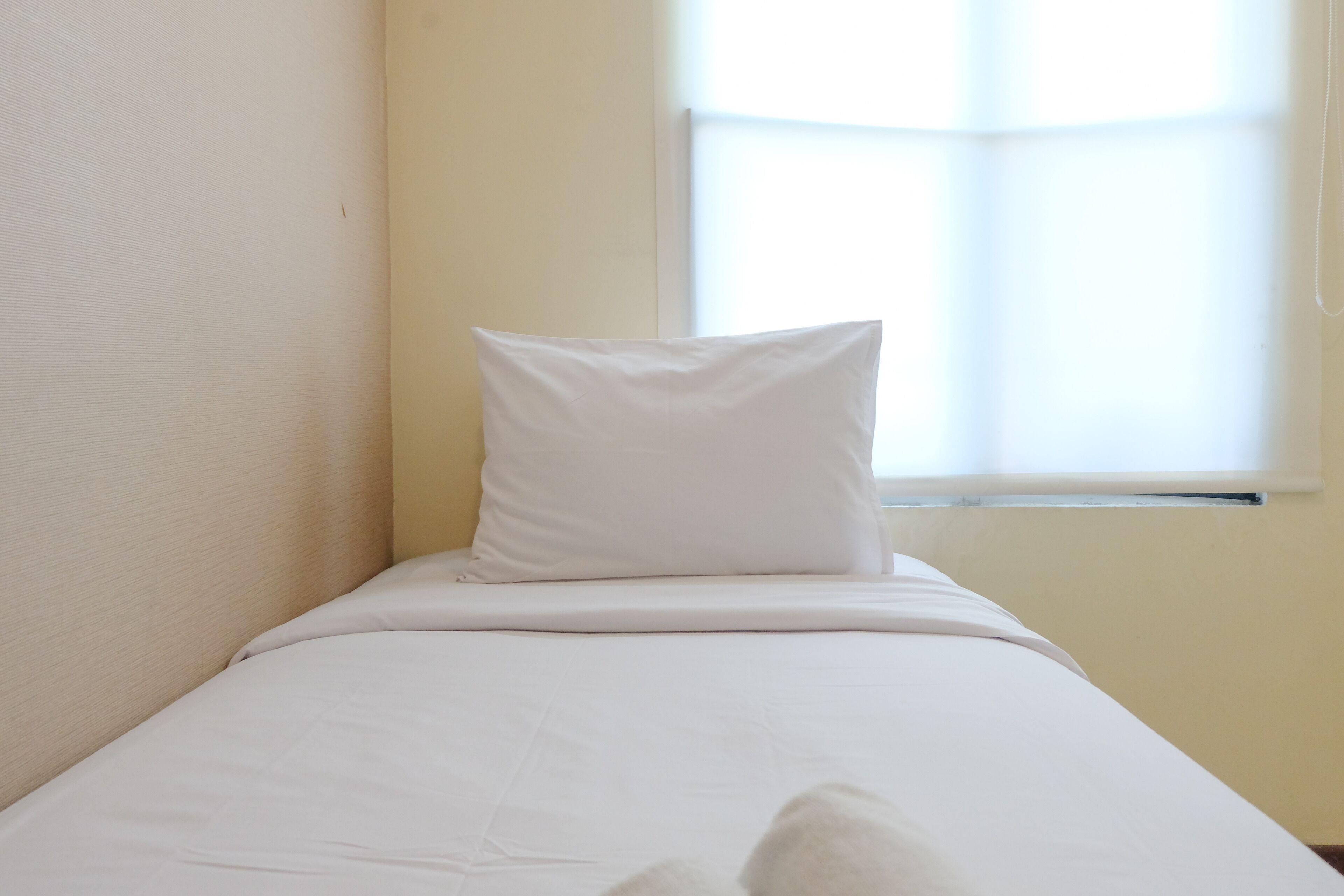 Una cama individual con ropa de cama blanca en una habitación con una pared lisa y una ventana.