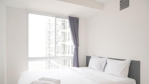 Ein ordentlich bezogenes Bett mit weißen Leinen und einem grauen Kopfteil, ein Fenster mit Blick auf Gebäude und ein grauer Vorhang.