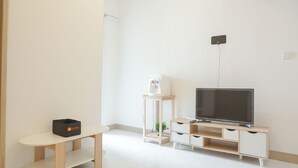 Ein schlicht eingerichteter Raum mit Fernseher auf einem Holzgestell, einem kleinen Tisch mit einer Kiste und einer weißen Wand.