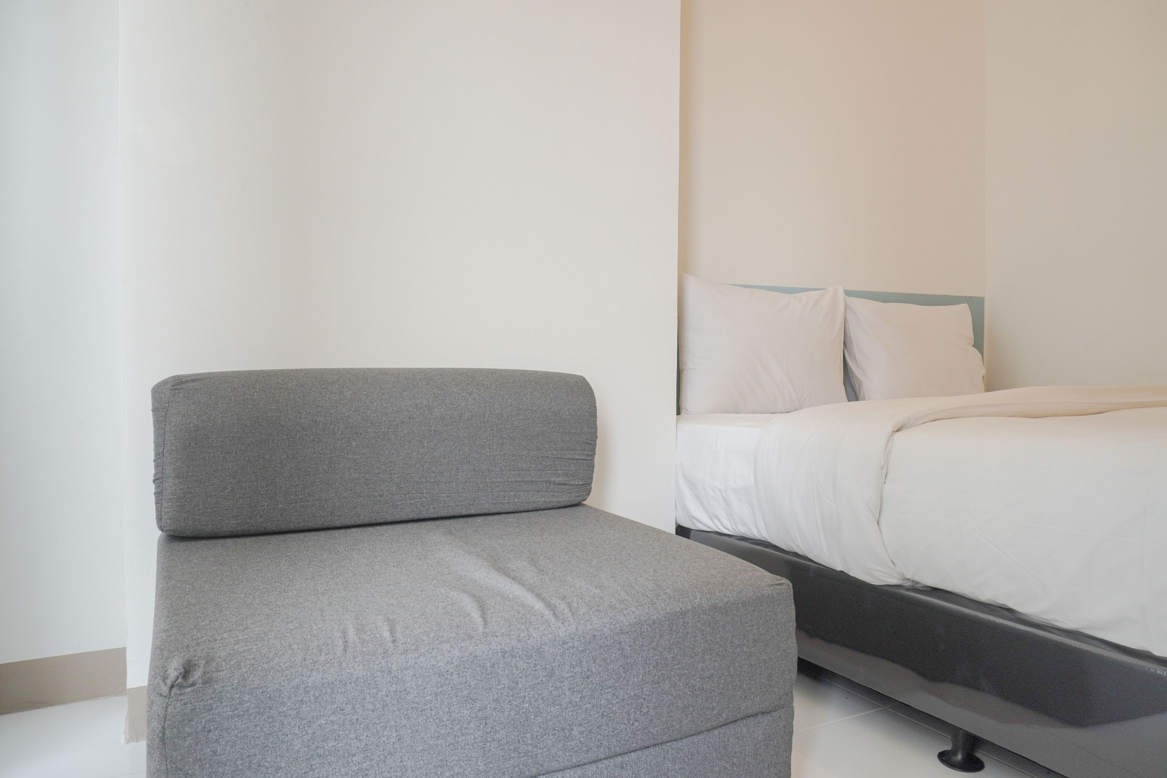 A modern bedroom with a grey upholstered chair, a bed with white bedding, and a plain wall.