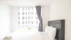 A neatly made bed with white linens, a grey curtain, and a view of a building through the window.