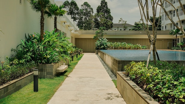A pathway leading to a pool surrounded by greenery and a building with a modern design. - Nice And Homey 2Br At Citra Living Apartment (Jakarta)