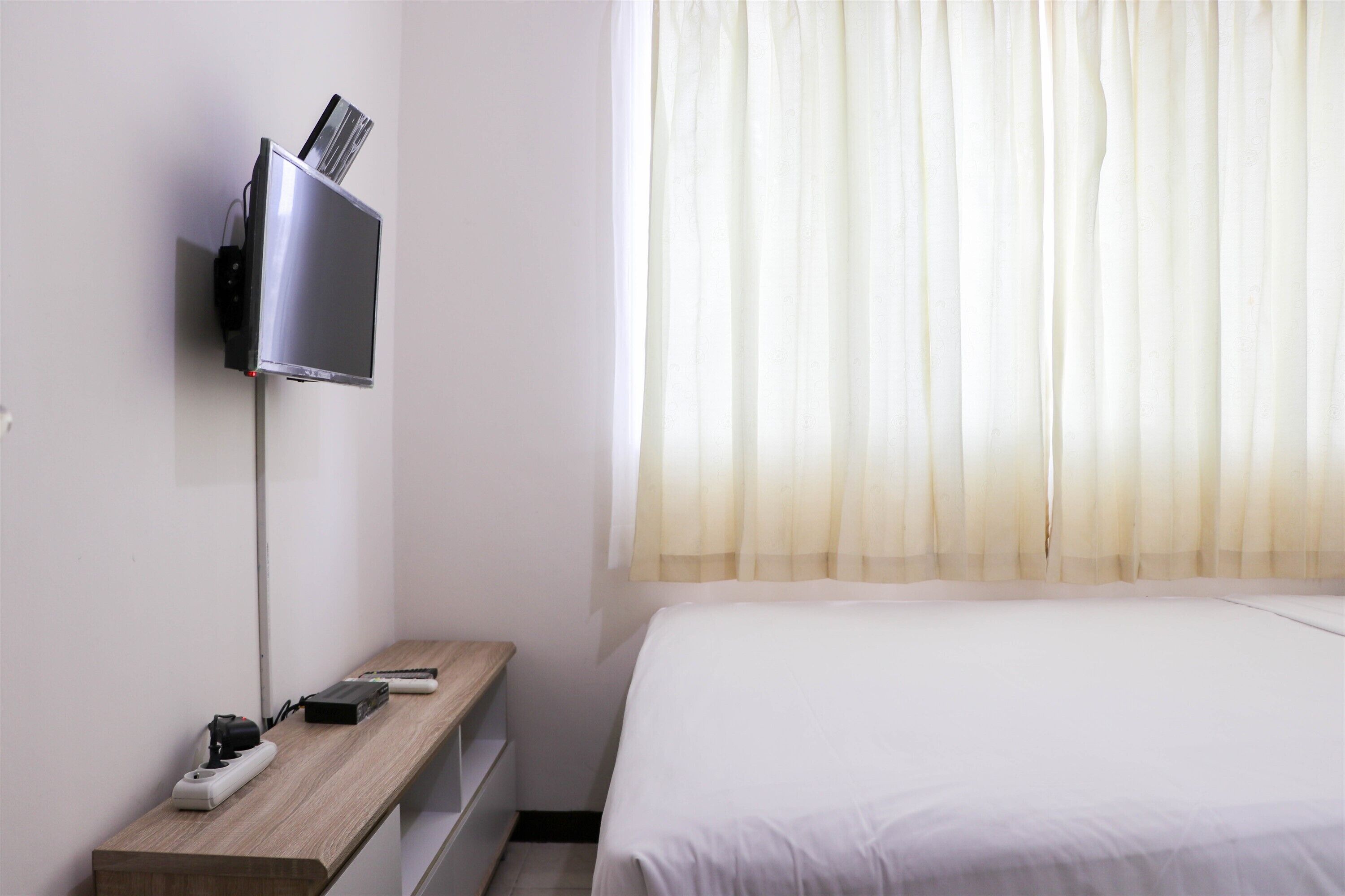 A hotel room with a bed, a TV mounted on the wall, a wooden nightstand, and a window with sheer curtains.