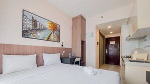 A hotel room with a bed, a desk, a chair, a painting, and a kitchenette. - Comfy And Best Choice Studio At Sky House Bsd Apartment (Pagedangan)