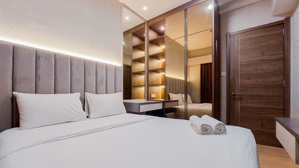 A modern hotel room with a large bed, a bedside table, a built-in wardrobe, and a mirror. - Spacious And Nice 2Br At Sky House Bsd Apartment (Pagedangan)