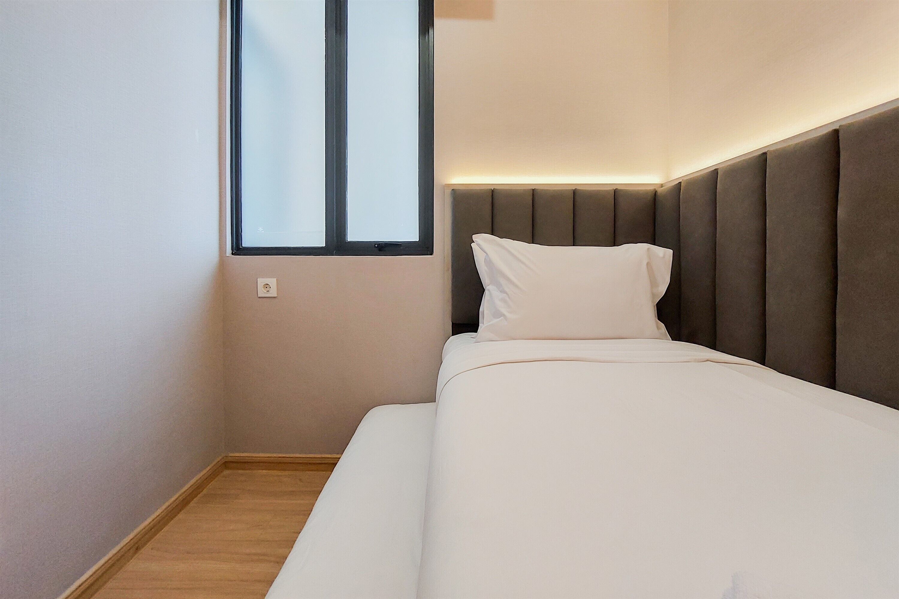 Ein ordentlich bezogenes Bett mit Kopfstütze, ein Fenster mit Jalousien und ein Lichtschalter an der Wand.