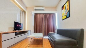 A modern living room with a grey sofa, wooden floor, a TV on a wooden stand, and an air conditioning unit above the curtains. - Spacious And Nice 2Br At Sky House Bsd Apartment (Pagedangan)