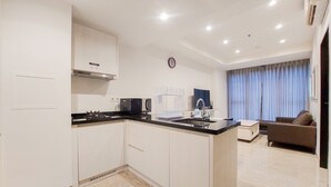 A modern kitchen with white cabinets, a black countertop, and a built-in stove. There is a dining area with a sofa and a window with curtains. - Homey And Tranquil 1Br Branz Bsd City Apartment (Pagedangan)