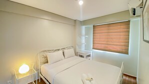 A neatly made bed with white linens and two rolled towels, a bedside table with a lamp, and a window with blinds. - Homey And Modern 1Br At Kebagusan City Apartment (Jakarta)