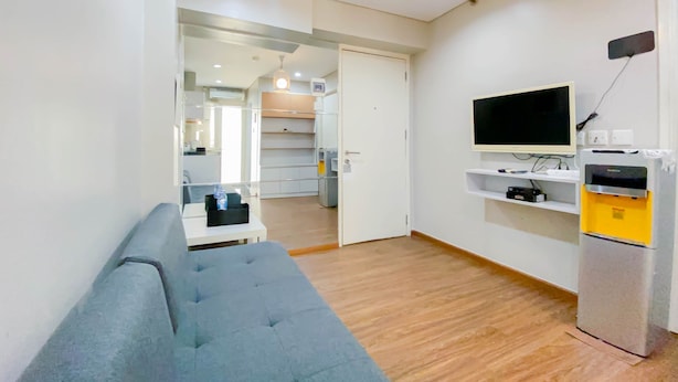 A modern living room with a grey sofa, a flat-screen TV mounted on the wall, and a built-in fridge. - Homey And Modern 1Br At Kebagusan City Apartment (Jakarta)