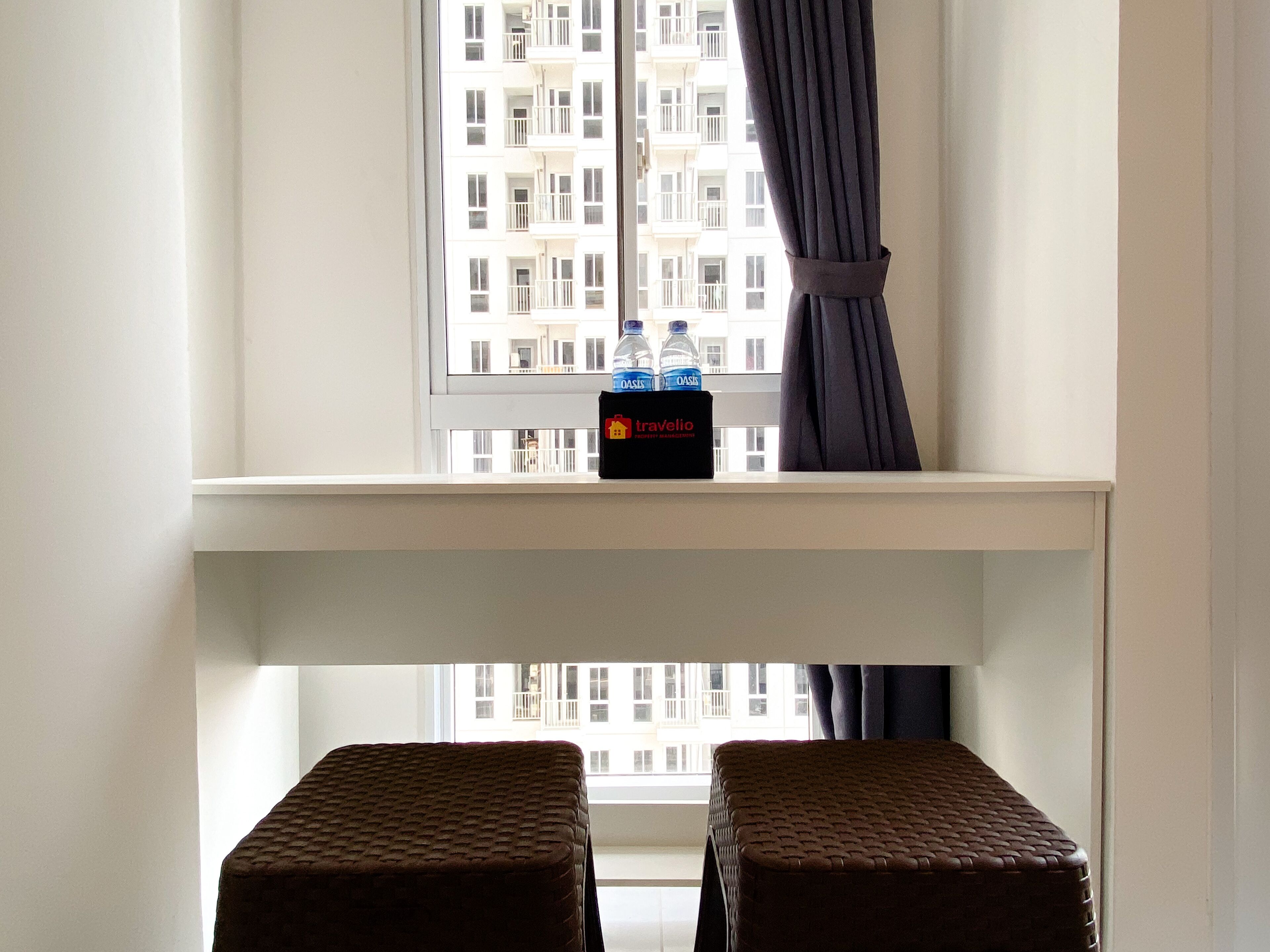 A room with a window overlooking a building, a small table with a box labeled 'travelio', and two wicker stools.