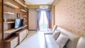 A living room with a TV on a wooden stand, a sofa, and a window with curtains. - Nice 2Br At Bogor Valley Apartment (Bogor)