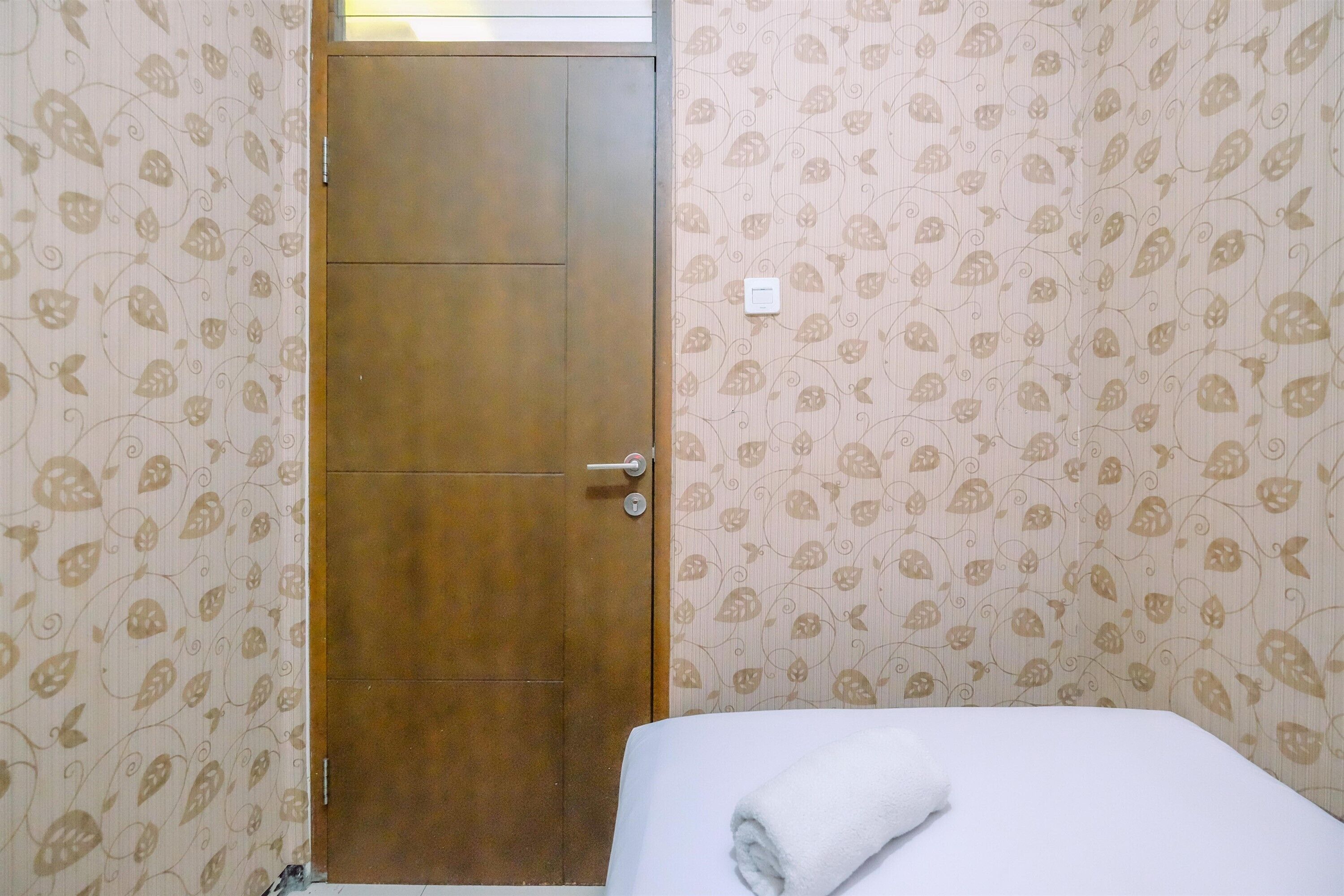 A hotel room with a wooden door, patterned wallpaper, and a neatly made bed with a white towel.