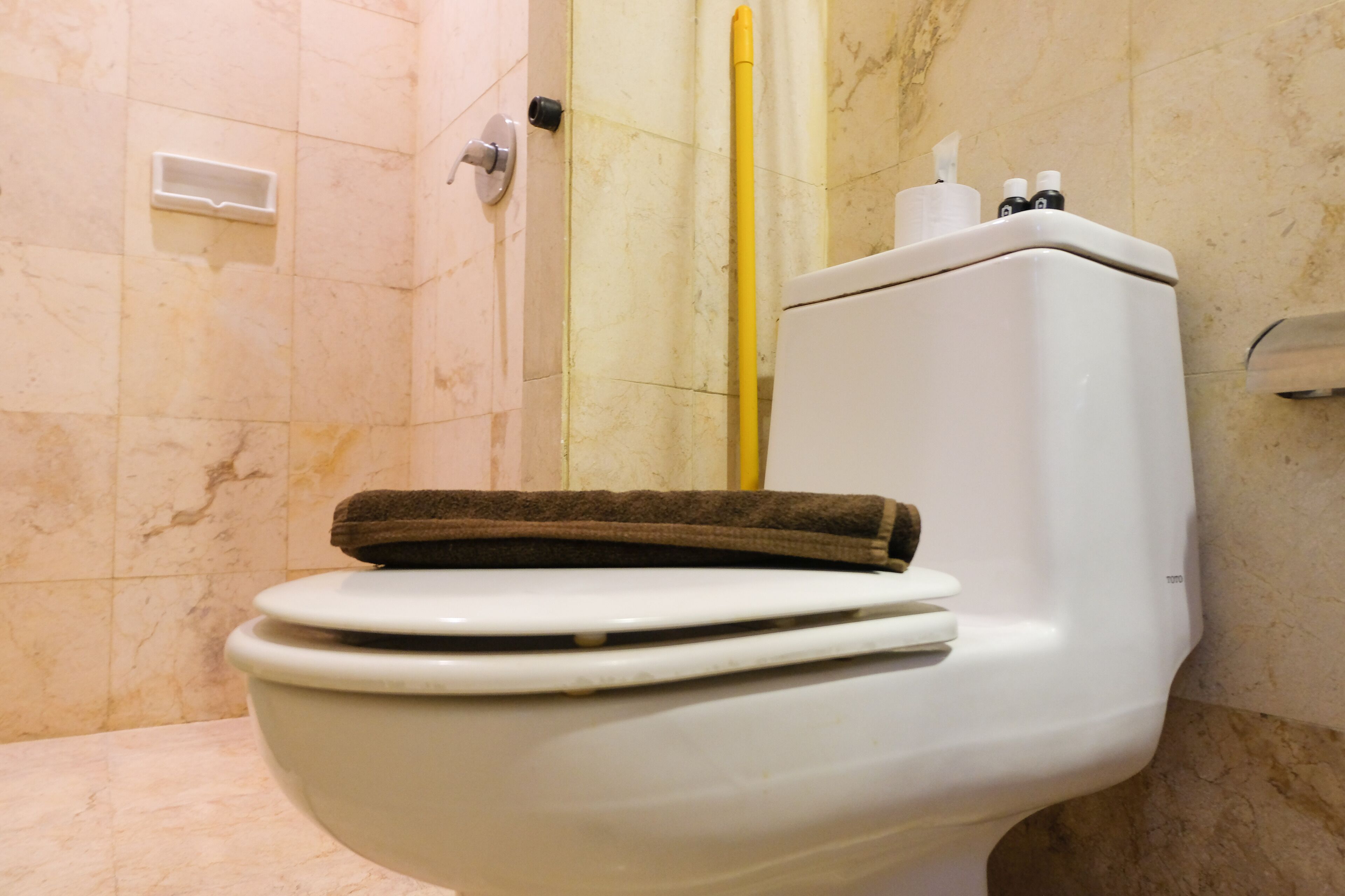 A white toilet with a brown towel on top, a yellow cleaning brush, and a soap dispenser in a bathroom with marble walls.