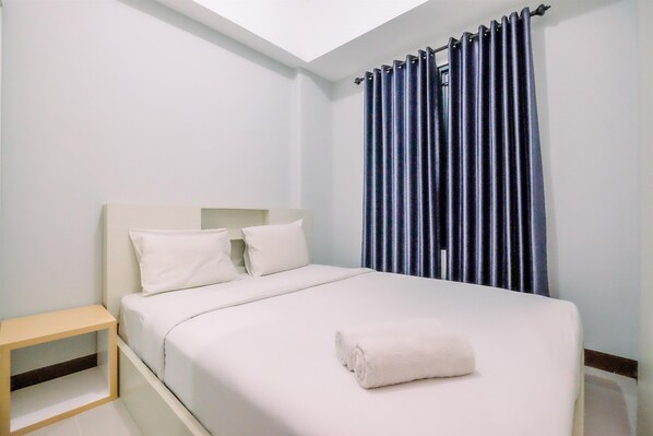 A neatly made bed with white bedding and pillows, a bedside table, and a window with blue curtains.