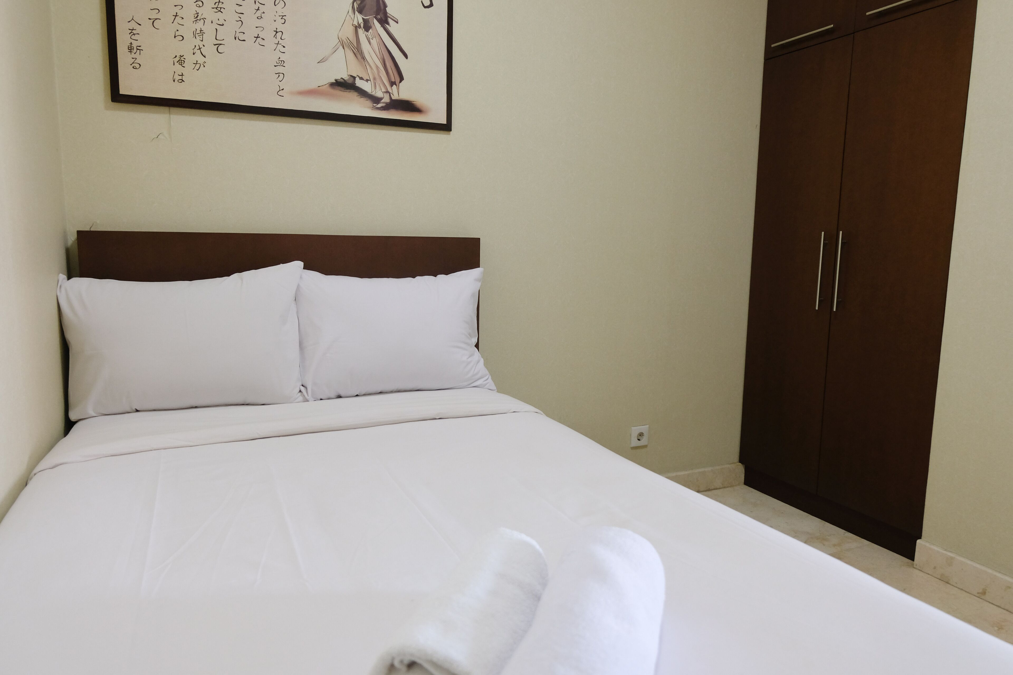 A neatly made bed with white linens and pillows, a wooden headboard, and a dark wooden wardrobe against a plain wall.