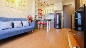 A living room with a sofa, a dining table with chairs, a fan, and a kitchen area with cabinets and appliances. - Modern And Good Deal 2Br At Transpark Cibubur Apartment (Depok)
