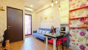 A compact living space with a sofa, a small dining table, a TV, and a kitchen area with a shelf. - Modern And Good Deal 2Br At Transpark Cibubur Apartment (Depok)
