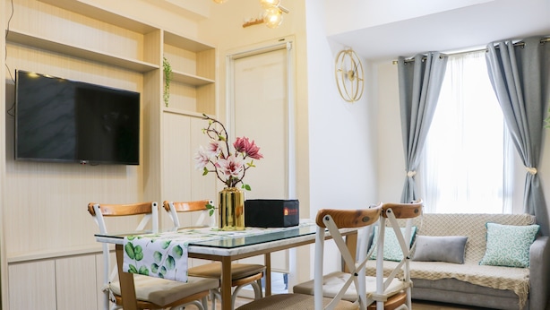 A living room with a TV mounted on the wall, a sofa with cushions, a dining table with chairs, and a vase with flowers. - Cozy Stay And Minimalist 1Br Tokyo Riverside Pik 2 Apartment (Teluknaga)