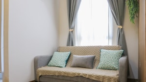 A grey sofa with a beige throw blanket and patterned cushions, positioned in front of a window with grey curtains tied back. - Cozy Stay And Minimalist 1Br Tokyo Riverside Pik 2 Apartment (Teluknaga)