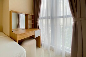 A hotel room with a bed, a wooden desk with a mirror, a small chair, and sheer curtains. - Well Furnished And Comfortable 2Br Apartment Gold Coast (Jakarta)