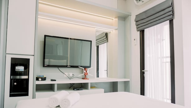 A modern hotel room with a large bed, a flat-screen TV mounted above the bed, a minibar, and a window with curtains.