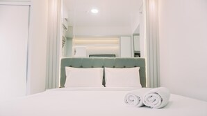 A neatly made bed with white linens and two rolled towels, a green upholstered headboard, and a white wall with a mirror and a light fixture.