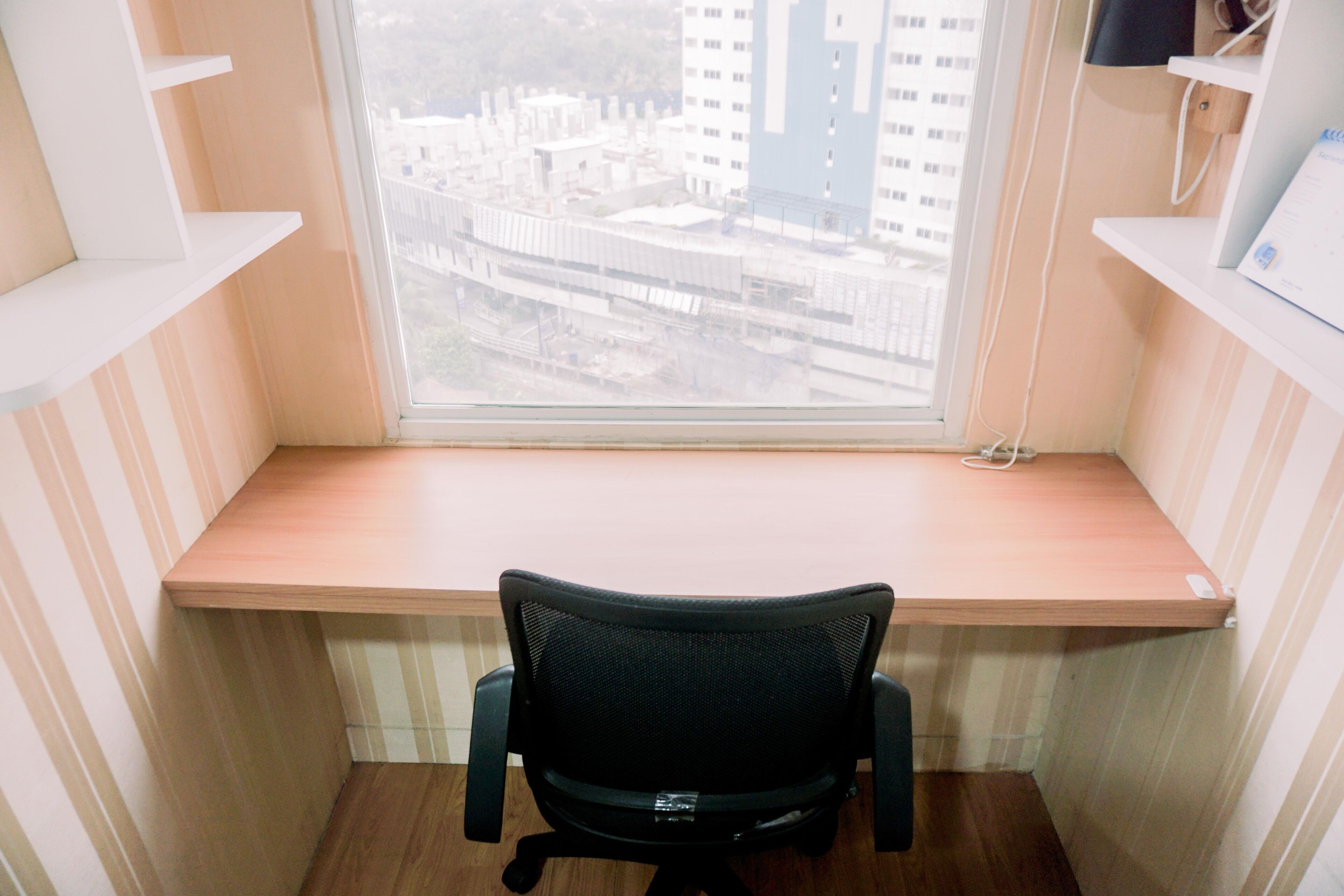 A corner desk with a chair, a window with a view of buildings, and a shelf with a computer monitor.