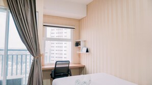 A hotel room with a bed, a desk with a chair, a window with curtains, and a view of buildings outside.