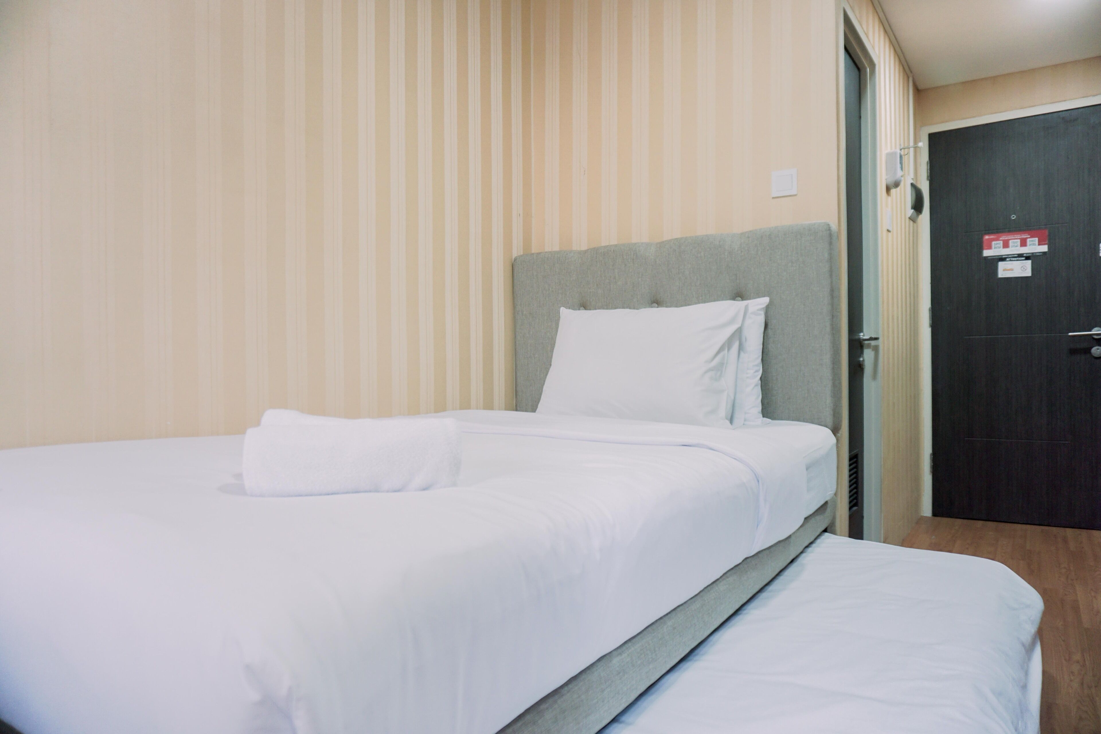 A hotel room with a single bed, a grey headboard, and a door with a red sign.