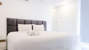 Un lit bien fait, avec du linge de lit blanc et deux oreillers, une tête de lit rembourrée de couleur grise et une armoire blanche avec un miroir.