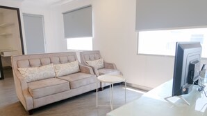 Un salon moderne avec un canapé beige, une petite table blanche et un bureau à plateau en verre sur lequel se trouve un écran d’ordinateur.