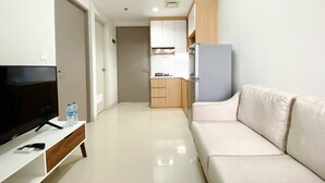 A modern living room with a flat-screen TV, a sofa, and a kitchen area with wooden cabinets and a refrigerator. - Great Deal And Pleasurable 2Br Vasanta Innopark Apartment (Cikarang)