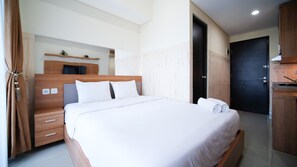 A modern hotel room with a wooden bed, a small bedside table, a built-in shelf with plants, and a dark door. - Strategic And Homey Studio At Tamasari Papilio Apartment (Surabaya)