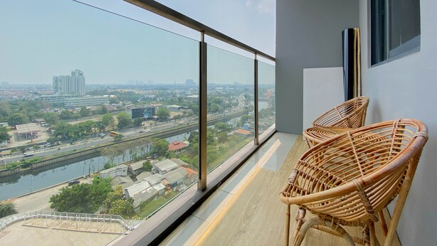 Een balkon met uitzicht op een rivier en gebouwen, met twee rieten stoelen en een glazen hekwerk.