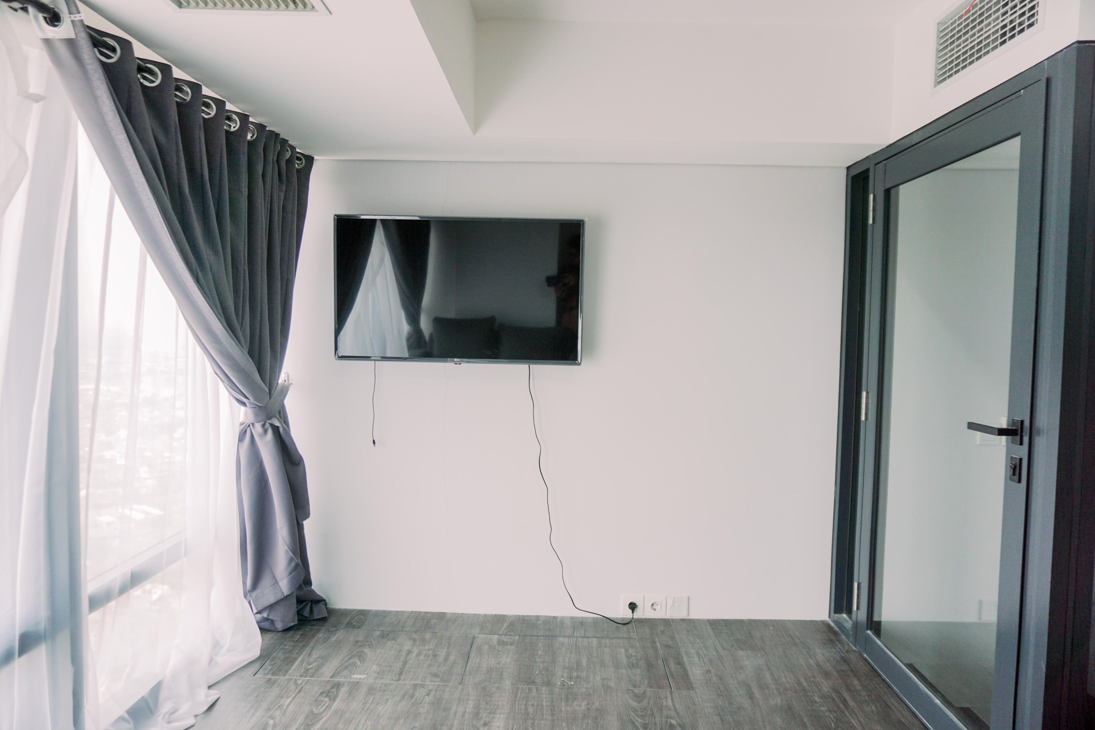 A room with a flat-screen TV mounted on the wall, a sliding glass door, and a window with sheer curtains.