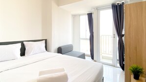 A hotel room with a large bed, a small armchair, a wooden wardrobe, and a balcony with curtains. - Furnished Studio For 3 Pax With Sofa Bed At Tokyo Riverside Pik 2 Apartment (Teluknaga)