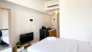 A hotel room with a bed, a TV on a wooden stand, a small plant, a mirror, a wardrobe, a desk, a refrigerator, a sink, and an air conditioning unit. - Furnished Studio For 3 Pax With Sofa Bed At Tokyo Riverside Pik 2 Apartment (Teluknaga)
