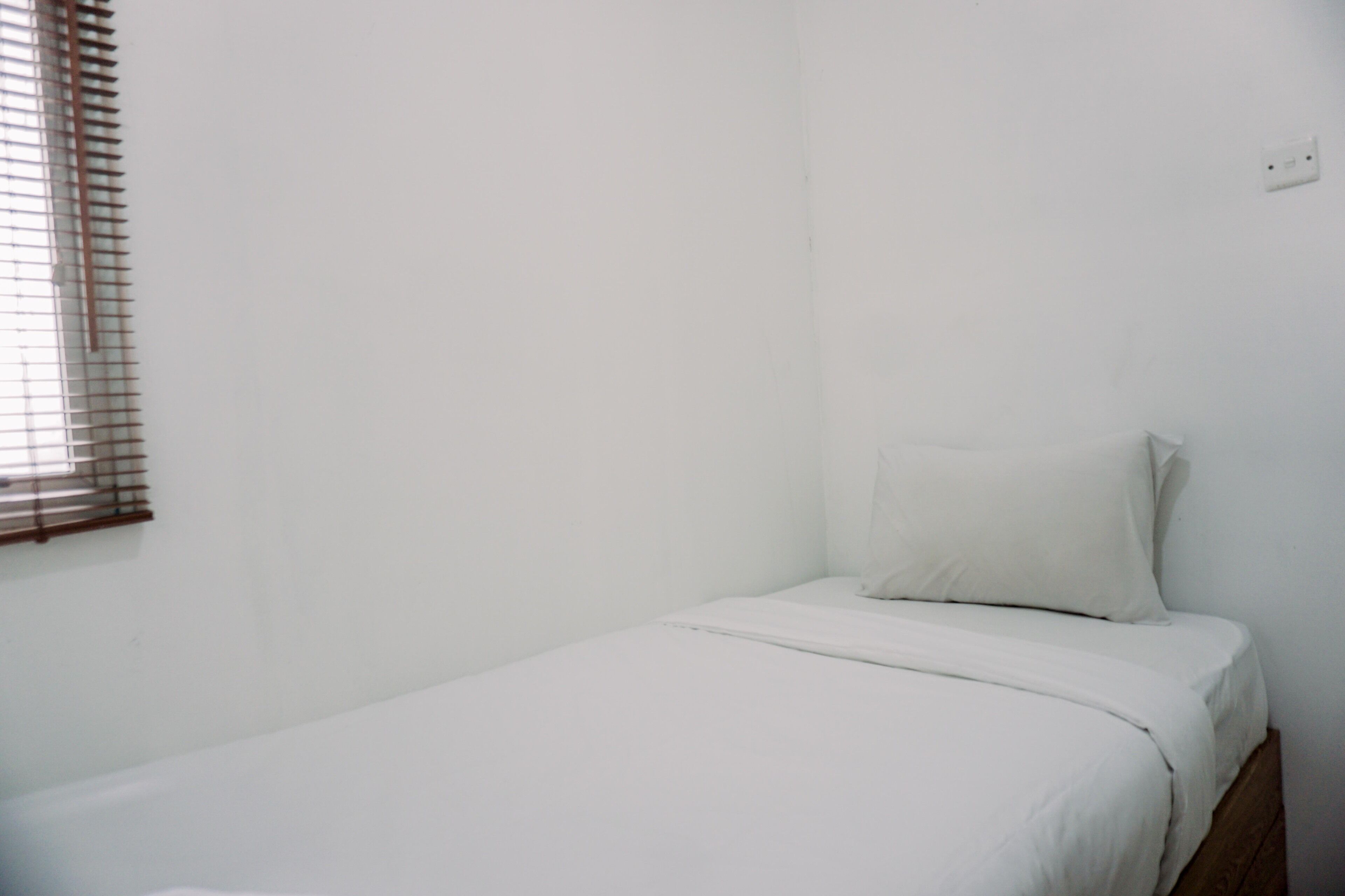 Ein kleines Zimmer mit einem Einzelbett, weißer Bettwäsche und einem Fenster mit Jalousien.