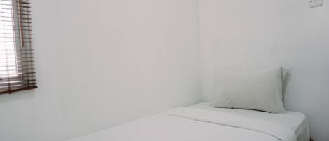Ein kleines Zimmer mit einem Einzelbett, weißer Bettwäsche und einem Fenster mit Jalousien.
