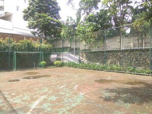 Una cancha de tenis exterior con red, rodeada por una cerca de malla metálica y follaje.