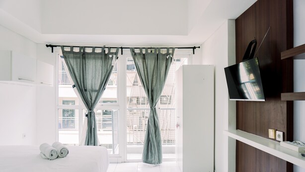 A hotel room with a bed, a TV mounted on the wall, and a balcony with curtains. - Homey And Good Deal Studio Casa De Parco Apartment (Pagedangan)