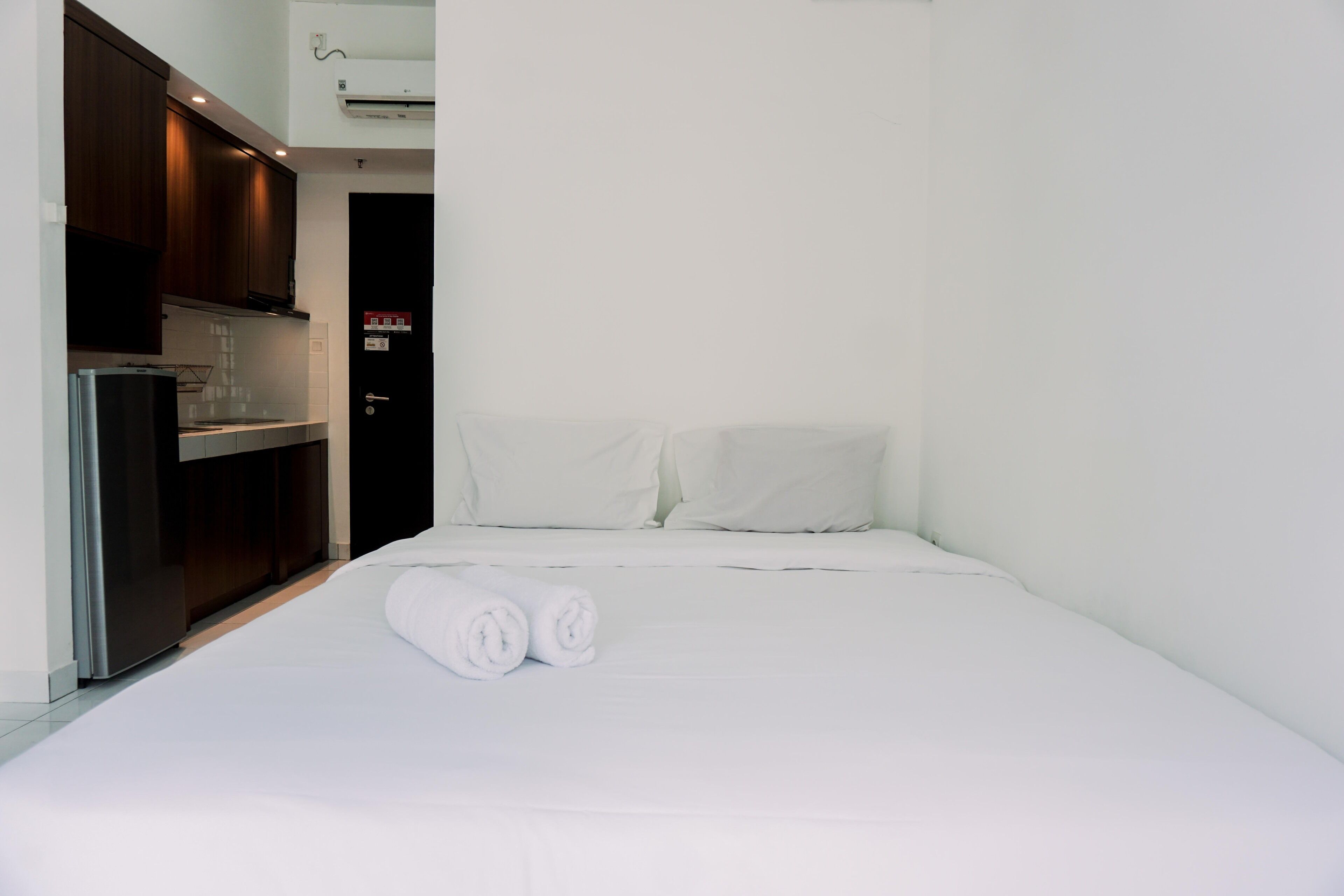 A small, neatly made bed with white linens and two rolled towels, a dark wooden wardrobe, and a small kitchenette with a refrigerator and microwave.