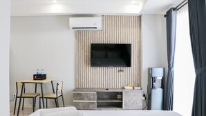 A hotel room with a bed, a TV mounted on a wooden panel wall, a desk with chairs, and a water dispenser. - Spacious And Homey Studio At Daan Mogot City Apartment (Jakarta)