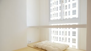Une chambre minimaliste avec un lit superposé, une fenêtre donnant sur un immeuble et un plancher en bois.