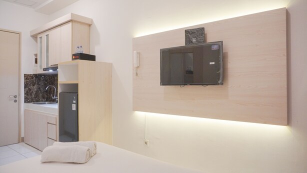 A modern hotel room with a kitchen area, a flat-screen TV mounted on a wooden panel, and a neatly folded towel on the bed. - Minimalist And Comfy Studio At Tokyo Riverside Pik 2 Apartment (Teluknaga)