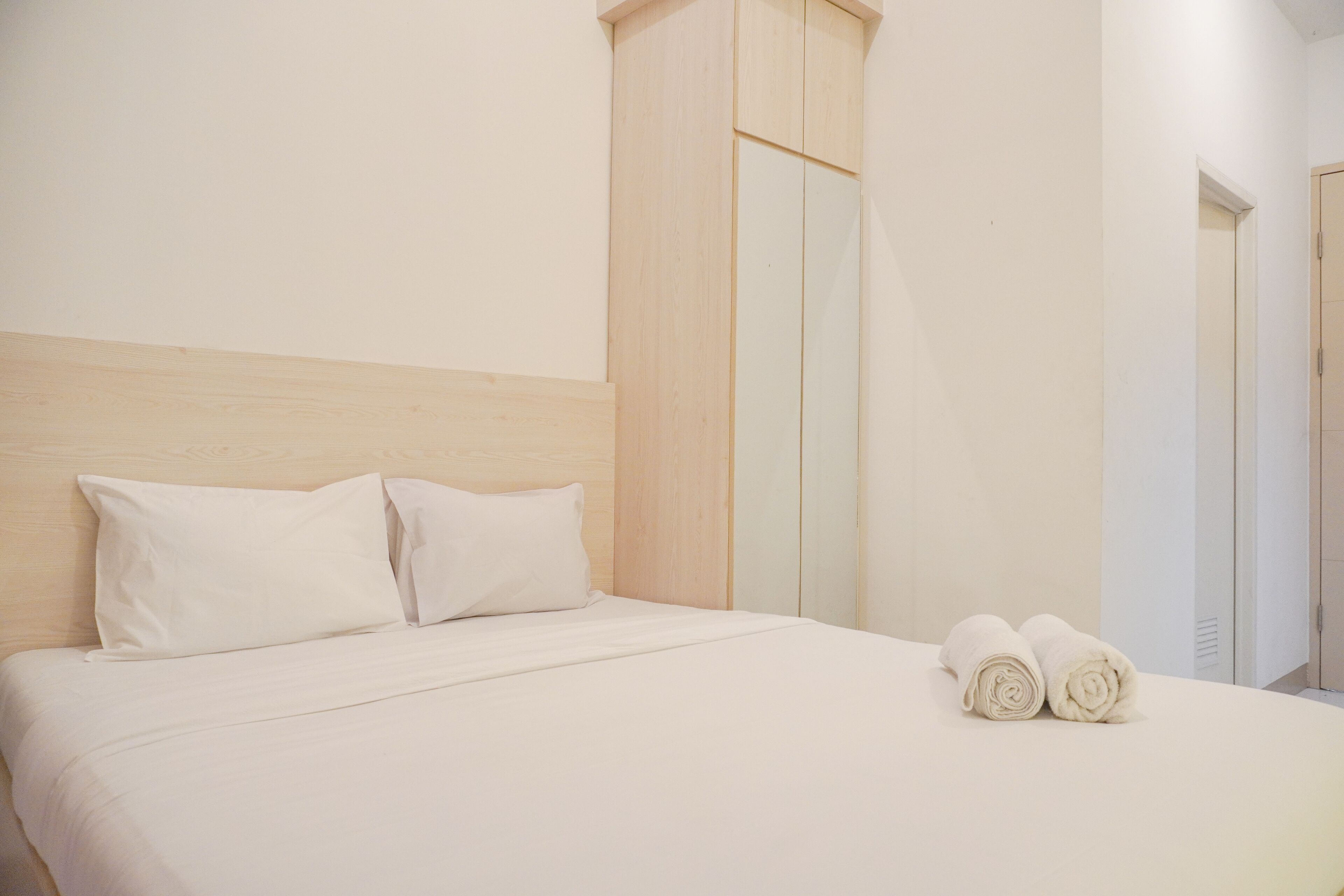 A neatly made bed with two pillows and two rolled towels, a wooden headboard, and a mirror on the wall.