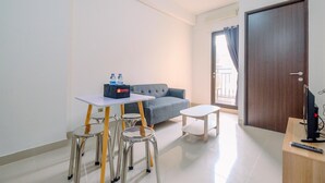 A modern living room with a grey sofa, a small white table, and a dining area with metal chairs. A TV and remote are on a wooden stand. - Wonderful 2Br At Transpark Cibubur Apartment (Bekasi)
